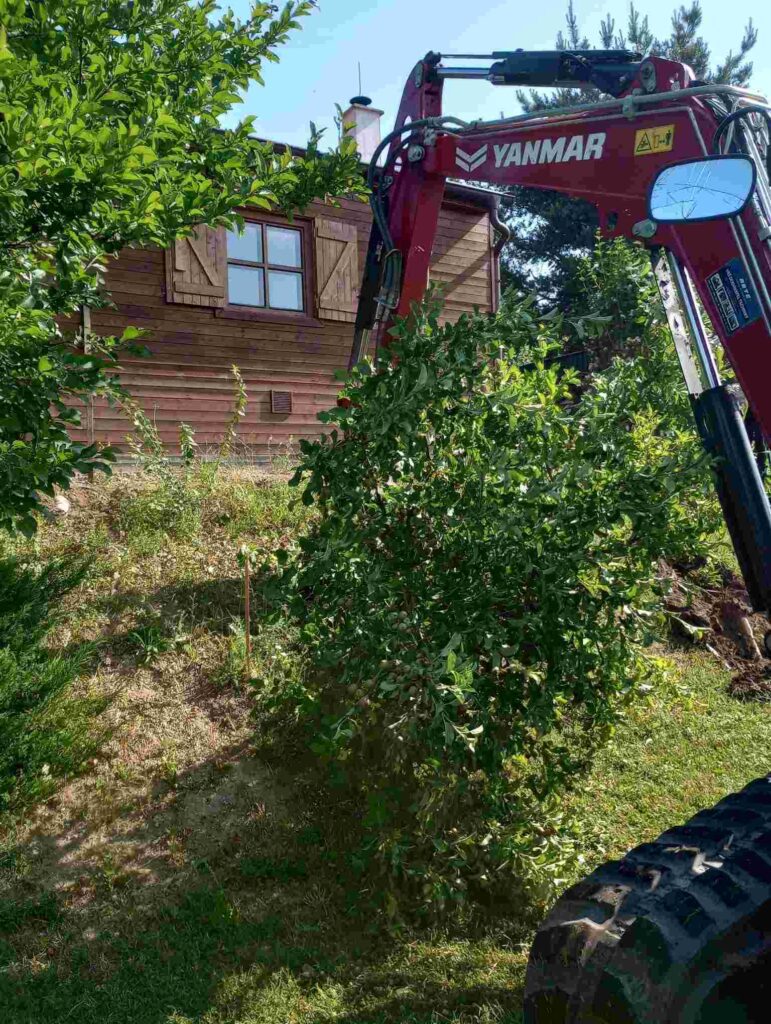 Bagr se připravuje na výkop pro instalaci plastového sklepu pod dřevěnou chatou. Jako precizní chirurg přesně zamířil svým ramenem ke svahu a chystá se vyhloubit důležitou díru. Vzácný pohled na synergii moderní technologie a tradiční architektury, která se spojuje ve prospěch pohodlí a úložného prostoru. Tato fotografie zachycuje začátek fascinujícího procesu, který oživí místo a přinese nový rozměr pobytu na chatě."